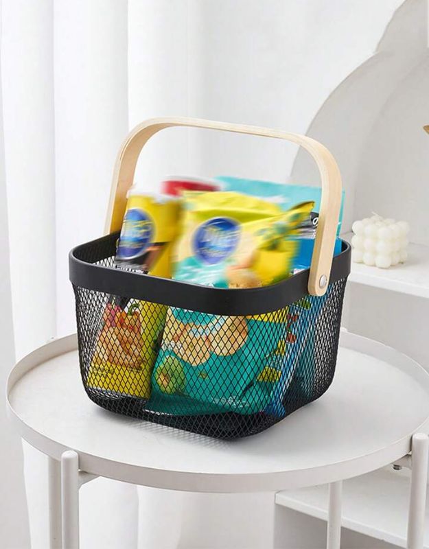A black metal mesh basket with a wooden handle filled with colorful items, placed on a white table.